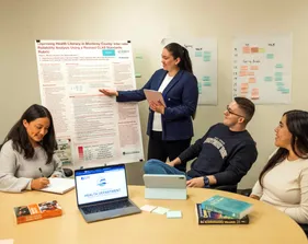 A student presenting a poster of work to three other students who are sitting at a table listening and taking notes.