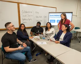 Five students sitting at a table listening while one of them is speaking to the professor who is standing smiling