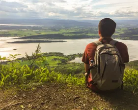 A boy with a backpack is sitting and looking at nature.