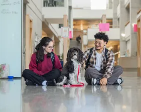 Two students sit on a library hallway floor petting a therapy dog between them.