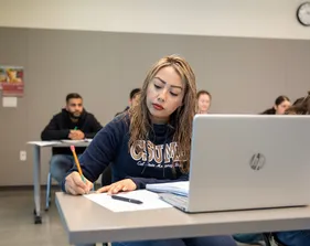 A student takes notes during a classroom session while using a laptop, demonstrating focus and active learning.