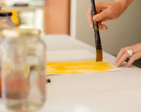A close-up of hands using a paintbrush to apply bright yellow paint onto paper, with jars of paint and water blurred in the foreground.