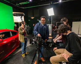 CSUMB Cinematic Arts students work together on a film set in a studio, adjusting a professional camera while others handle audio, lighting, and a red car scene in front of a green screen.