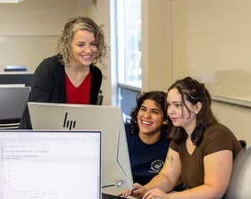 A faculty member works with two students at computer stations in a classroom highlighting mentoring, collaboration and hands-on learning at Cal State �鶹��.