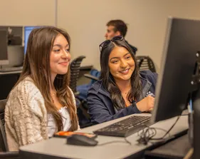 Two girl are smiling and looking atorientation, student collaboration, computer lab, business administration, peer mentoring, campus learning, academic support, new student experience a computer screen