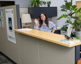 A woman stands behind a desk, smiling warmly and waving hello.