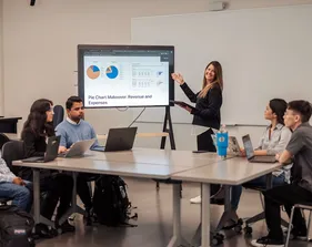 A faculty member presents financial charts on a screen to a group of students seated around a table with laptops during an accounting class.