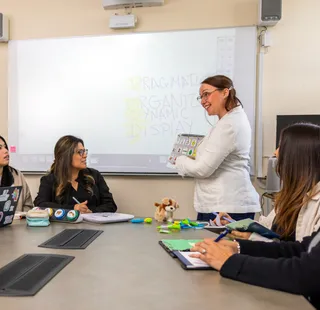 Professor lecturing her students in front of a whiteboard while they are sitting at a table.