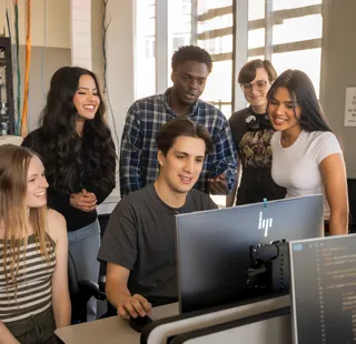 A group of students looking at a students desktop while they work on it