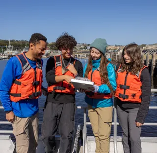 A group of four students standing on a boat with life jackets on looking at a clipboard