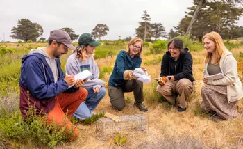 A group of four AES students and a professor kneeling in the grass while the professor holds a squirrel
