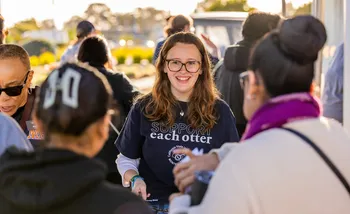A smiling Cal State Monterey Bay staff member wearing a “Support each otter” shirt greets admitted students and families during Admitted Otter Day, engaging in conversation at an outdoor campus welcome event.