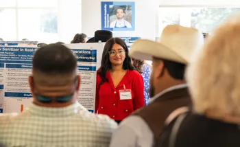 Student in a red shirt presents her agricultural research poster to an audience at CSUMB’s AGPS Summer showcase. Attendees stand nearby and ask questions.