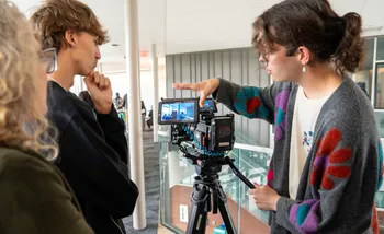 Person demonstrating a camera setup on a tripod while explaining framing to two others indoors.