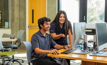 A smiling student sitting down at a desk using a computer, with a school counselor peering over their shoulder and pointing at the computer screen with a smile