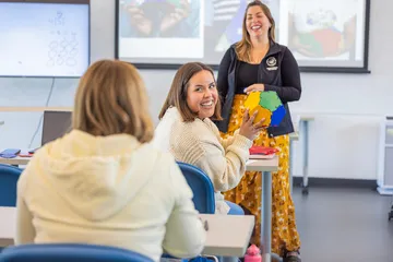 A student smiles while holding a colorful geometric model as a professor leads an engaging classroom discussion.