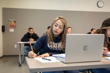 A student takes notes during a classroom session while using a laptop, demonstrating focus and active learning.