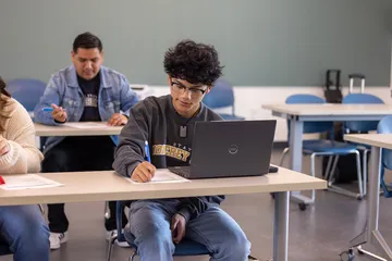 A student works independently in a classroom, using a laptop while completing an assignment.