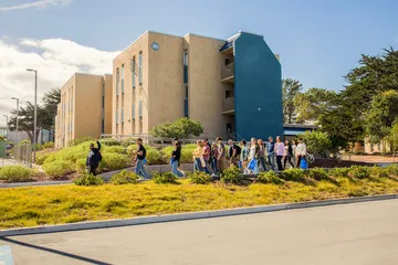 A group of students walks together across campus near residential buildings, enjoying a sunny day in a welcoming community setting