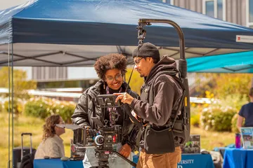 Two CSUMB Cinematic Arts students review footage on a camera monitor while operating a professional film camera rig under a canopy during an outdoor campus event.