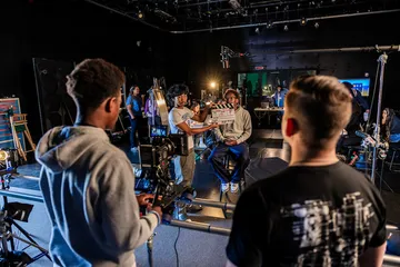 Students in the Cinematic Arts and Technology program at Cal State Monterey Bay film a scene in a professional studio, with one student operating a camera while another holds a clapperboard and crew members prepare lighting and sound equipment.