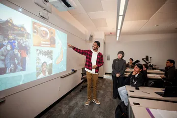 A student stands at the front of a classroom pointing to a projected presentation featuring photos and Japanese cultural imagery, while classmates and an instructor watch from their seats.