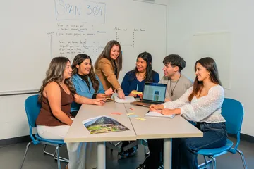 An instructor and six CSUMB students collaborate around a table in a Spanish literature classroom. The instructor leans in to guide discussion while students share color-coded note cards, consult a laptop displaying Spanish-language text, and take notes. A whiteboard behind them reads “SPAN 304” with handwritten Spanish analysis, highlighting guided and collaborative learning in the Spanish langua