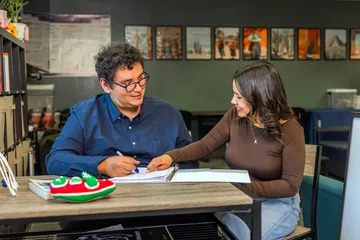 A CSUMB instructor and a student sit together at a table during a one-on-one Spanish language learning session. The instructor writes in a notebook while the student points and smiles, engaging in conversation. Bookshelves and cultural imagery in the background create a supportive and focused learning environment.