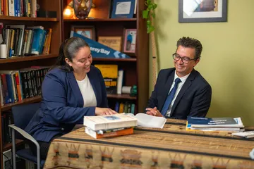 Student explaining something to a professor who is listening with books in the background.