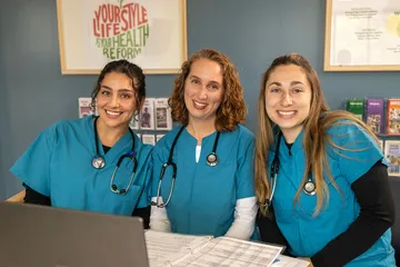 A group of three nursing students in scrubs with stethoscopes around their necks sitting down and smiling at the camera