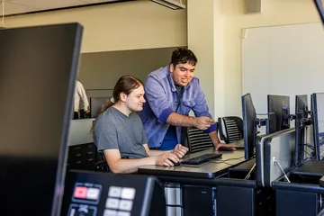 A computer science student collaborates with a classmate in a computer lab, reviewing code and working at desktop stations, highlighting mentoring, collaboration and hands-on learning at Cal State Monterey Bay.