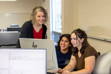 A faculty member works with two students at computer stations in a classroom highlighting mentoring, collaboration and hands-on learning at Cal State �鶹��.