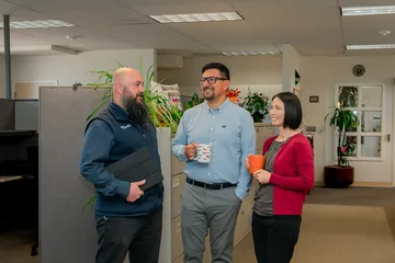 Three CSUMB HR staff members share a friendly conversation near a cubicle partition lined with plants and an “Inclusive Space” sign, smiling and holding coffee mugs and a tablet.