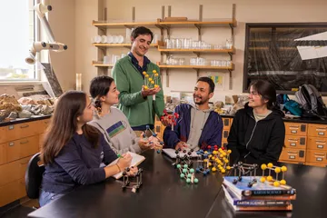 Instructor holding a molecular model while students sit around a table examining and discussing models in a chemistry lab.