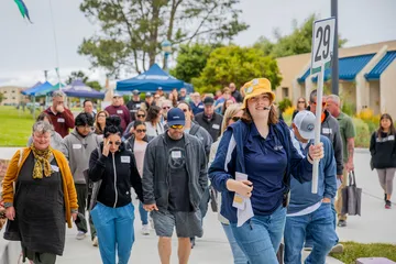 A student ambassador leads a campus tour group along a sidewalk at Cal State �鶹��, holding a route sign while prospective students, families and guests walk together during an orientation event.