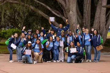 A group of Cal State �鶹�� orientation leaders pose outdoors during a transfer student orientation event, smiling, waving and holding schedules as they welcome new students to campus.