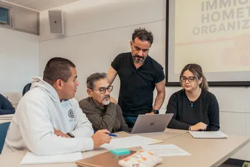A faculty member stands with a small group of students seated at a table, reviewing material on a laptop during a classroom discussion on immigrant hometown organizations.
