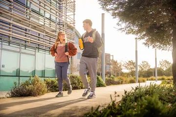 Two ���ӽ紫ý college students walk and chat on a sunny campus path, carrying backpacks and a water bottle near a modern building.