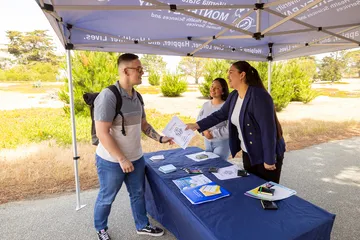 Two women manning a table and handing out a flyer to a man that is standing in front of the booth.