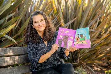 A woman sitting on a bench outdoors smiles while holding open a colorful children’s book featuring illustrations of raised fists and people eating tacos, with text in both English and Spanish.