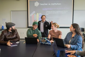 Professor gestures while speaking with a group of smiling students using laptops around a classroom table