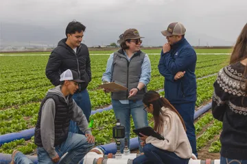 Five students in a field, checking a pipe and recording data on their notebooks.