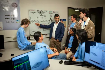 Six students looking at the professor teach while he is pointing at a whiteboard.