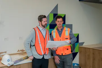 Two male staff members in safety vests review construction documents near a CSUMB Dining Common wall with abstract green and blue designs, discussing site updates.