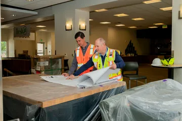 Two male facilities team members review blueprints on a wrapped countertop at CSUMB’s dining commons, surrounded by construction materials and signage.
