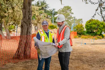 wo CSUMB facilities professionals review blueprints at an outdoor construction site, standing under trees near orange safety fencing and discussing the project layout.