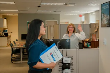 An HR staff member waves goodbye to a smiling employee holding onboarding documents marked “E-Verify” while standing in an open and friendly office space.