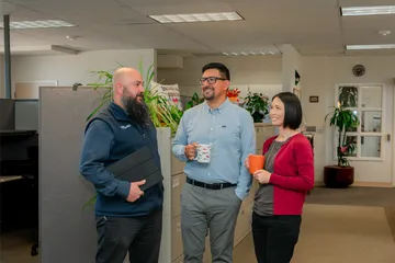 Three CSUMB HR staff members share a friendly conversation near a cubicle partition lined with plants and an “Inclusive Space” sign, smiling and holding coffee mugs and a tablet.