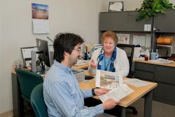 An HR specialist at CSUMB meets with a staff member in a private office, discussing employee benefits with printed documents while seated at a desk.