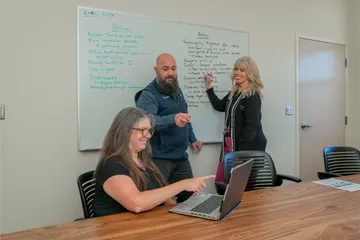 CSUMB HR staff collaborate during a “Glow & Grow” feedback session in a conference room, writing notes on a whiteboard while another team member takes notes on a laptop.